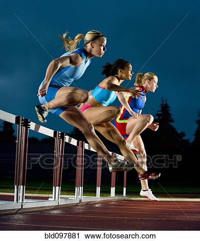 Stock Image - Runners jumping hurdles in race. Fotosearch