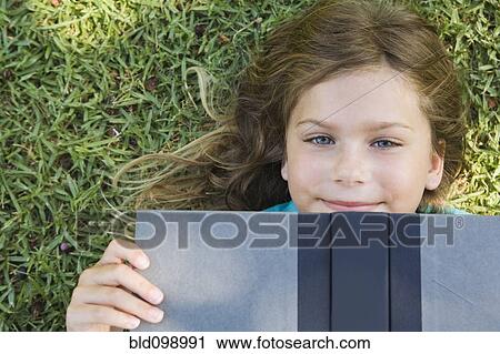 Stock Image - Smiling girl laying on ground reading book. Fotosearch