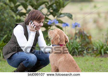Stock Photo - Smiling Hispanic woman talking on cell phone and petting dog. Fotosearch