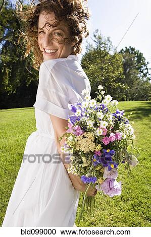 Stock Image - Smiling woman holding flowers in park. Fotosearch