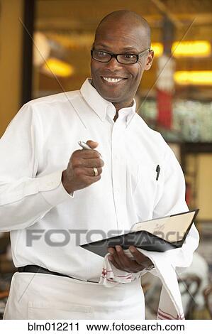 Stock Image - African American waiter holding order pad in restaurant. Fotosearch
