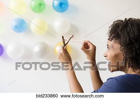 Stock Image - Black woman aiming slingshot at balloons on wall. Fotosearch