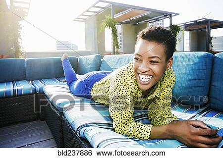 Stock Photograph - Black woman laying on rooftop sofa laughing. Fotosearch