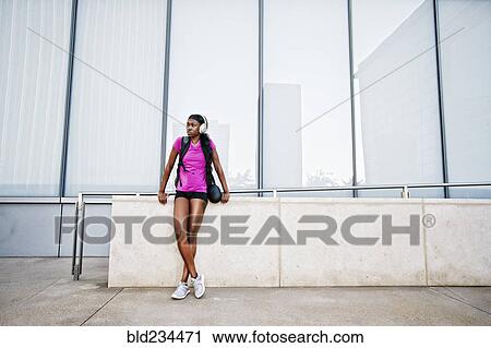 Stock Image - Black woman leaning on urban wall listening to headphones. Fotosearch