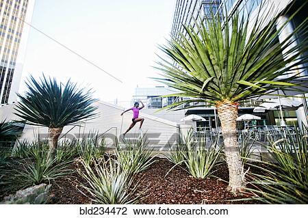 Stock Image - Black woman running and jumping on sidewalk near trees. Fotosearch