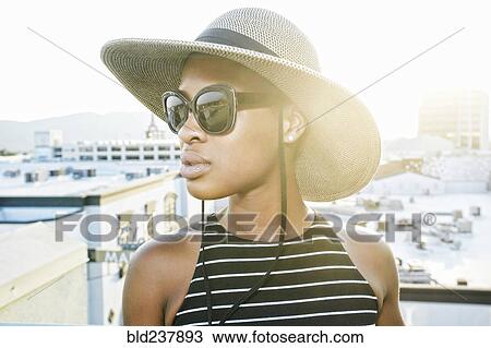 Stock Image - Black woman wearing sun hat and sunglasses on rooftop. Fotosearch