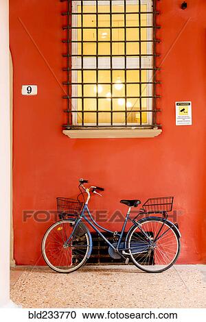 Stock Image - Blue bicycle leaning on orange wall under window. Fotosearch