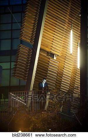 Stock Image - Businessman on balcony texting on cell phone at night. Fotosearch