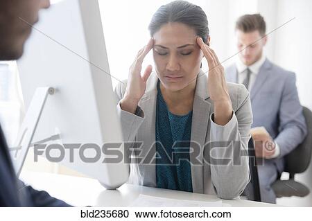 Stock Image - Businesswoman with headache rubbing temples in office. Fotosearch