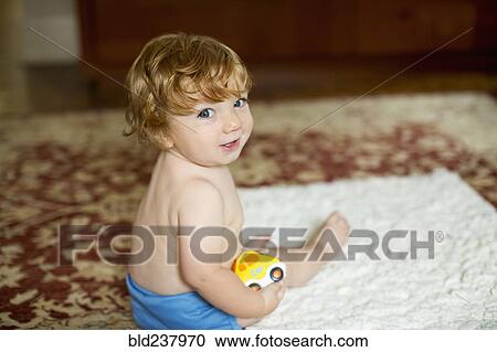 Stock Image - Caucasian baby boy sitting on rug holding toy car. Fotosearch