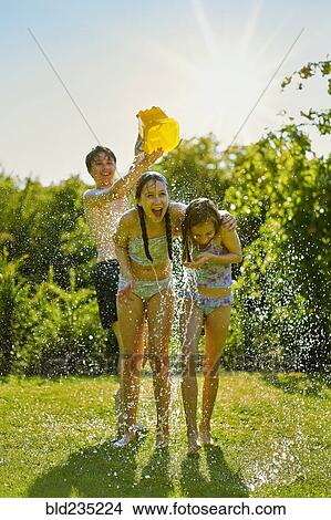 Picture - Caucasian boy dumping bucket of water on girls in backyard. Fotosearch