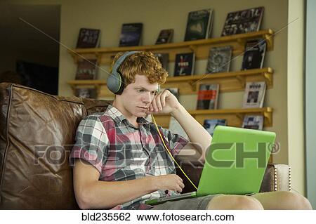 Stock Image - Caucasian boy sitting on sofa listening to laptop with headphones. Fotosearch