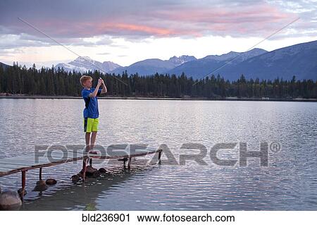 Stock Image - Caucasian boy standing on wooden dock photographing lake. Fotosearch
