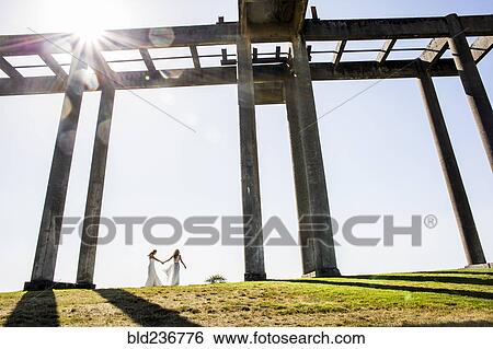 Stock Photograph - Caucasian brides holding hands walking under concrete structure. Fotosearch