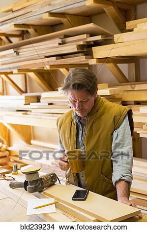 Picture - Caucasian carpenter drinking coffee and using cell phone in workshop. Fotosearch