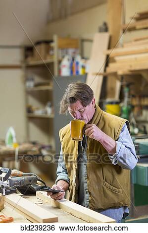 Stock Photograph - Caucasian carpenter drinking coffee and texting on cell phone in workshop. Fotosearch