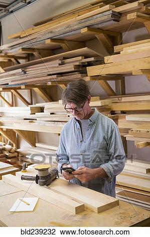 Stock Image - Caucasian carpenter texting on cell phone in workshop. Fotosearch