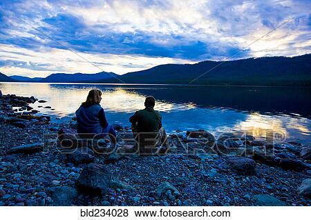 Stock Photo - Caucasian couple sitting on rocks at lake during sunset. Fotosearch