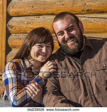 Stock Photograph - Caucasian couple smiling near log cabin wall. Fotosearch