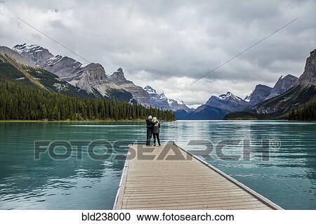 Stock Image - Caucasian couple standing at the end of dock on mountain lake. Fotosearch