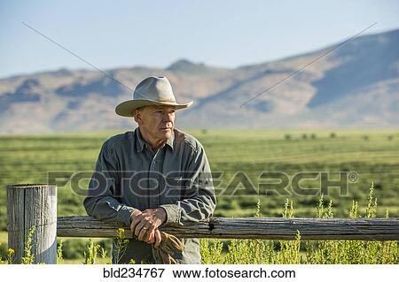 Stock Photo - Caucasian farmer holding gloves leaning on wooden fence. Fotosearch