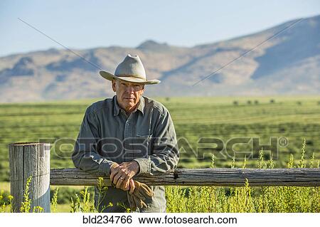 Stock Photograph - Caucasian farmer holding gloves leaning on wooden fence. Fotosearch