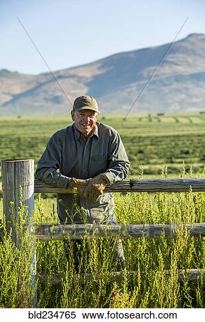 Stock Photography - Caucasian farmer wearing gloves leaning on wooden fence. Fotosearch