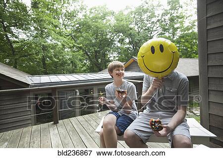 Stock Photo - Caucasian father and son eating donuts with smiley face balloon. Fotosearch