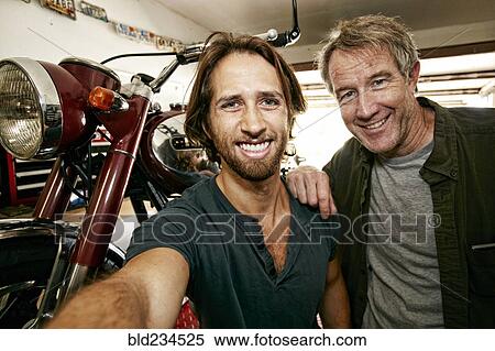 Stock Photography - Caucasian father and son posing for selfie with motorcycle in garage. Fotosearch