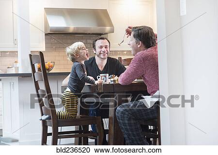 Stock Image - Caucasian father feeding son at table. Fotosearch