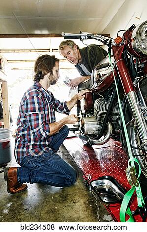 Stock Photo - Caucasian father watching son repairing motorcycle in garage. Fotosearch