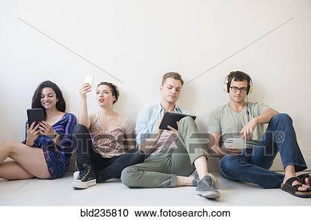 Stock Image - Caucasian friends sitting on floor using technology. Fotosearch