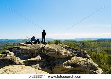Stock Image - Caucasian friends sitting on mountain rock admiring scenic view. Fotosearch