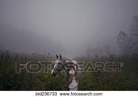 Stock Image - Caucasian girl petting horse in foggy field. Fotosearch