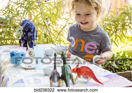 Stock Image - Caucasian girl playing with tea set and dinosaurs. Fotosearch