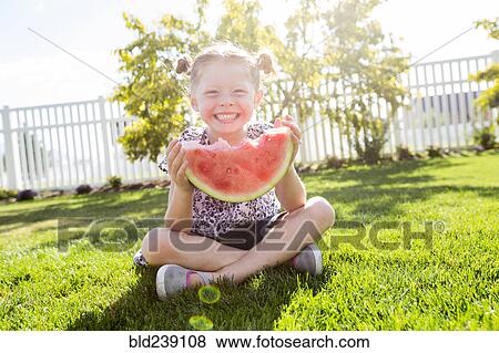 Stock Photo - Caucasian girl sitting in grass eating watermelon. Fotosearch