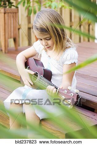 Stock Image - Caucasian girl sitting on patio playing ukulele. Fotosearch