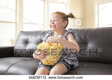 Stock Image - Caucasian girl sitting on sofa eating bowl of popcorn. Fotosearch