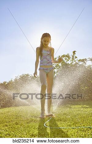 Picture - Caucasian girl standing over backyard sprinkler. Fotosearch