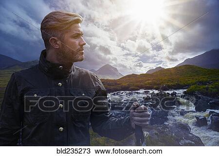 Stock Image - Caucasian hiker holding walking stick near river. Fotosearch