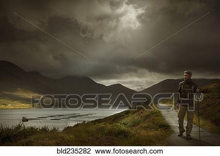 Stock Image - Caucasian hiker holding walking stick on path near river. Fotosearch