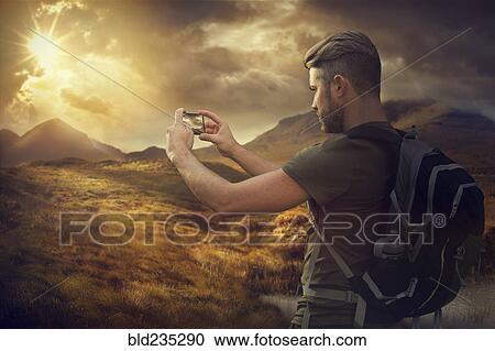 Stock Image - Caucasian hiker photographing mountain sunset with cell phone. Fotosearch