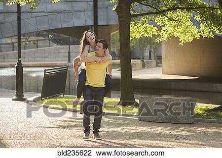 Stock Image - Caucasian man carrying girlfriend piggyback in park. Fotosearch