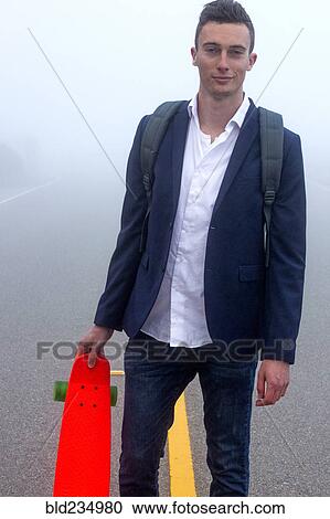 Stock Image - Caucasian man holding small red skateboard in fog. Fotosearch