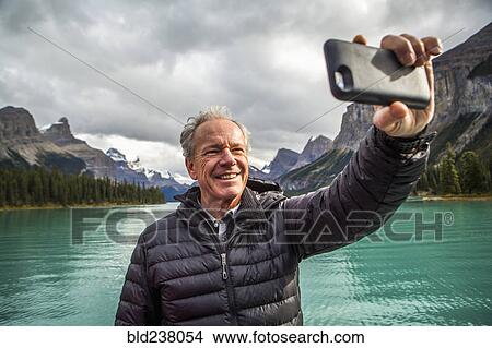 Picture - Caucasian man posing for cell phone selfie near mountain lake. Fotosearch