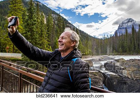 Stock Image - Caucasian man posing for cell phone selfie near mountain. Fotosearch