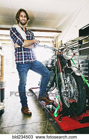 Stock Image - Caucasian man posing with motorcycle in garage. Fotosearch