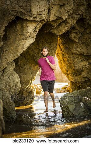 Stock Image - Caucasian man running under rock formation at beach. Fotosearch