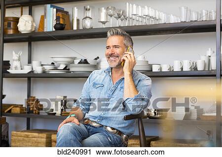 Stock Image - Caucasian man sitting in store talking on cell phone. Fotosearch