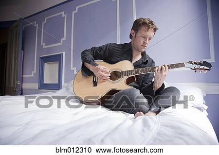 Stock Image - Caucasian man sitting on bed playing guitar. Fotosearch